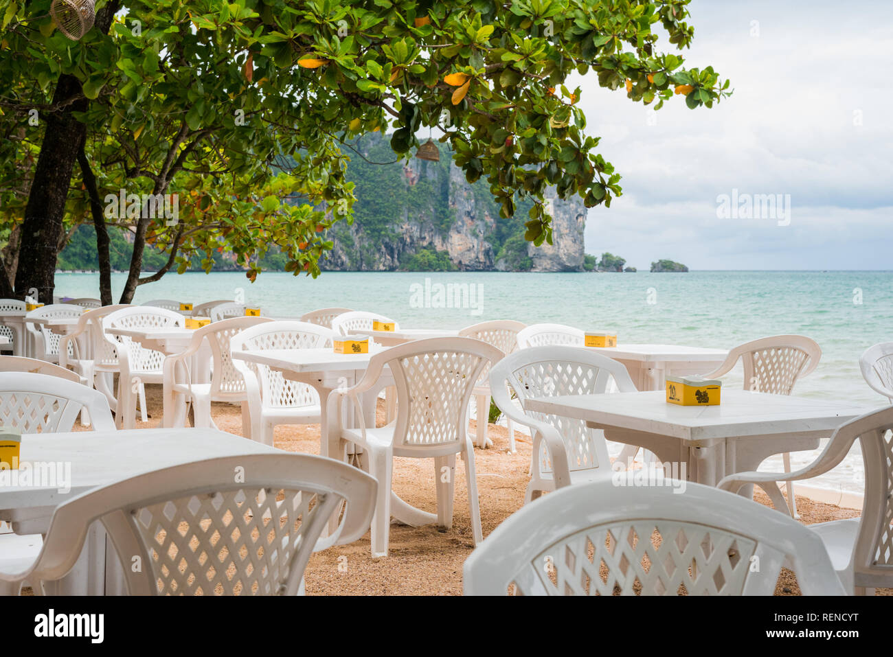 Ao Nang, Tailandia - July 2, 2018: tablas vacías de una cafetería al aire libre en la playa de Ao Nang contra el cielo nublado y el mar grisáceo. Foto de stock