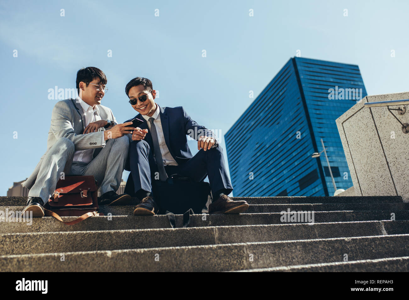 Dos socios de negocios sentados en las escaleras al aire libre mirando el teléfono móvil. hombre de negocios que muestra algo interesante a su compañero sentado en sta Foto de stock