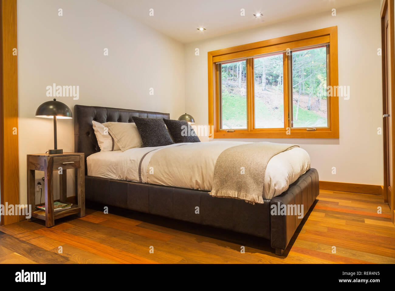 Cama king size con cabecera de panel de cuero marrón y mesas de madera con lámparas iluminadas en el dormitorio de invitados con piso de madera Ipe en casa de lujo. Foto de stock