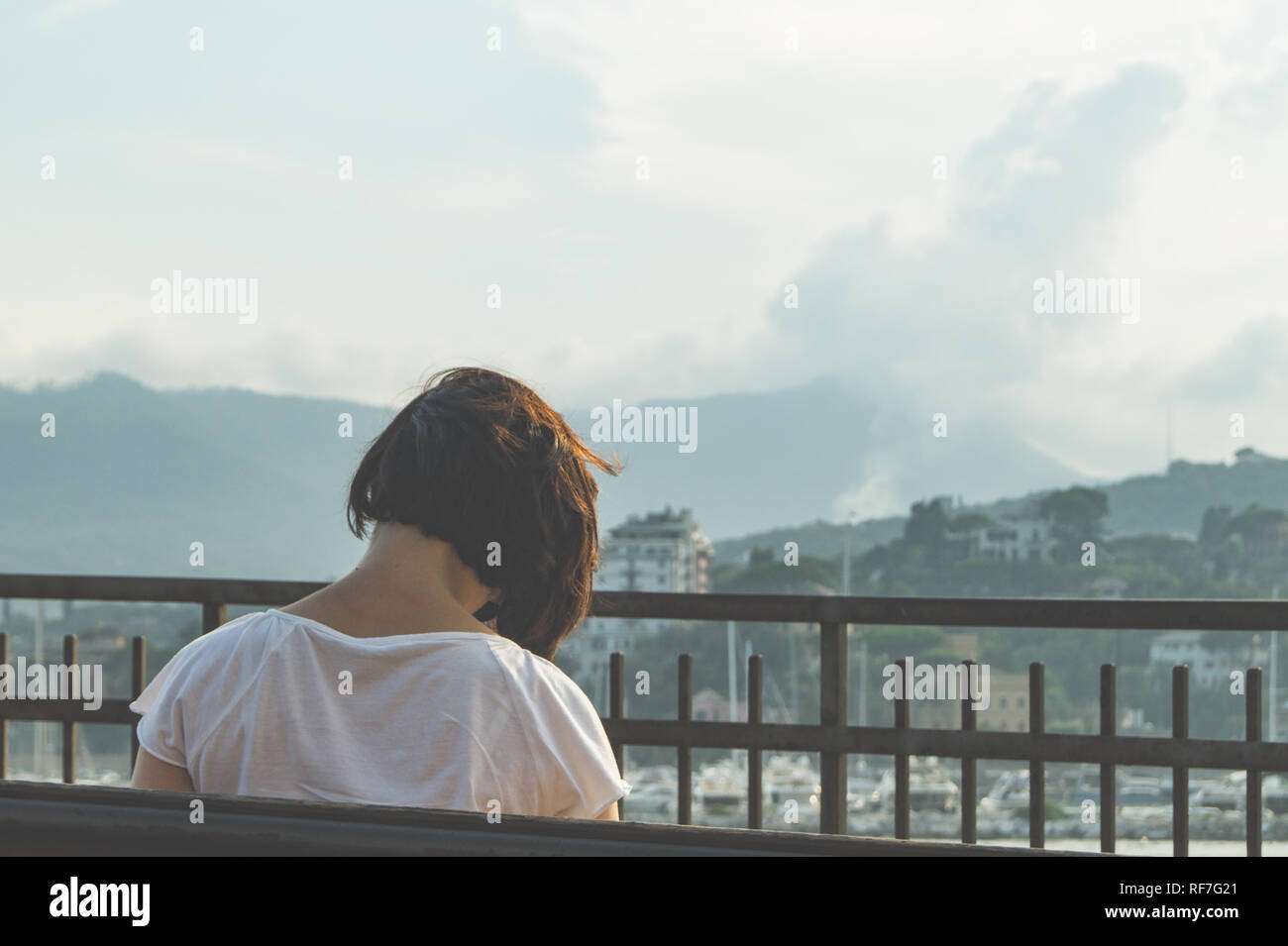 vista posterior de una mujer inclinando su cabeza mientras se sienta en un banco frente al paisaje Foto de stock