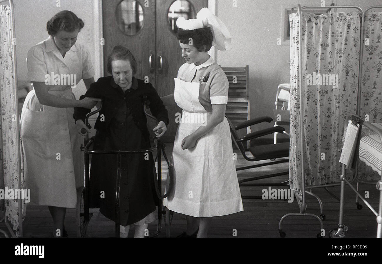 1950s, históricos, dos enfermeras ayudando a un herido lady paciente en una cátedra de movilidad moverse dentro de un hospital, Inglaterra, Reino Unido. Foto de stock