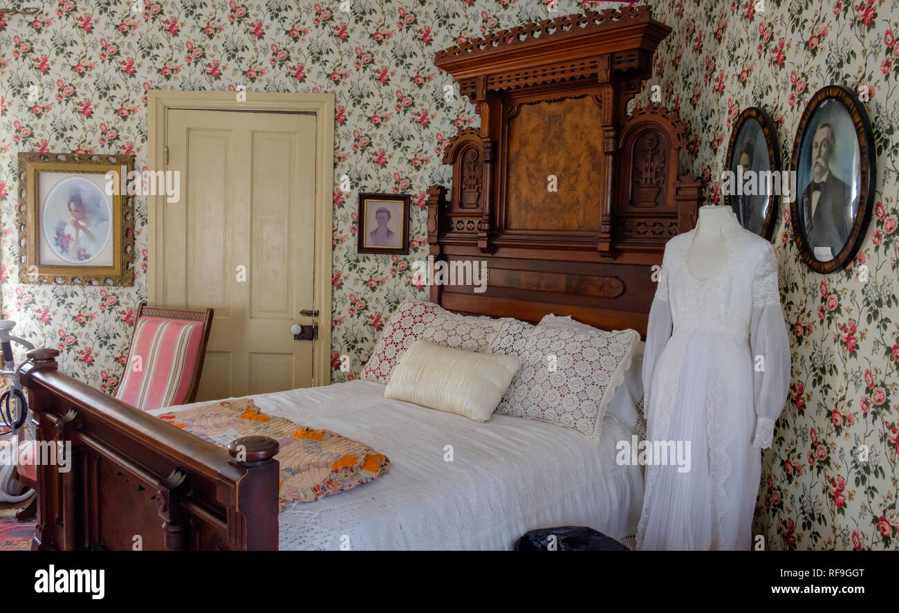 Antique interior del dormitorio con cama doble, cabecero de madera ornamentados y escalón, fundas de almohadas, encaje vestido de boda vintage, papel de pared con motivos florales, retratos enmarcados. Foto de stock