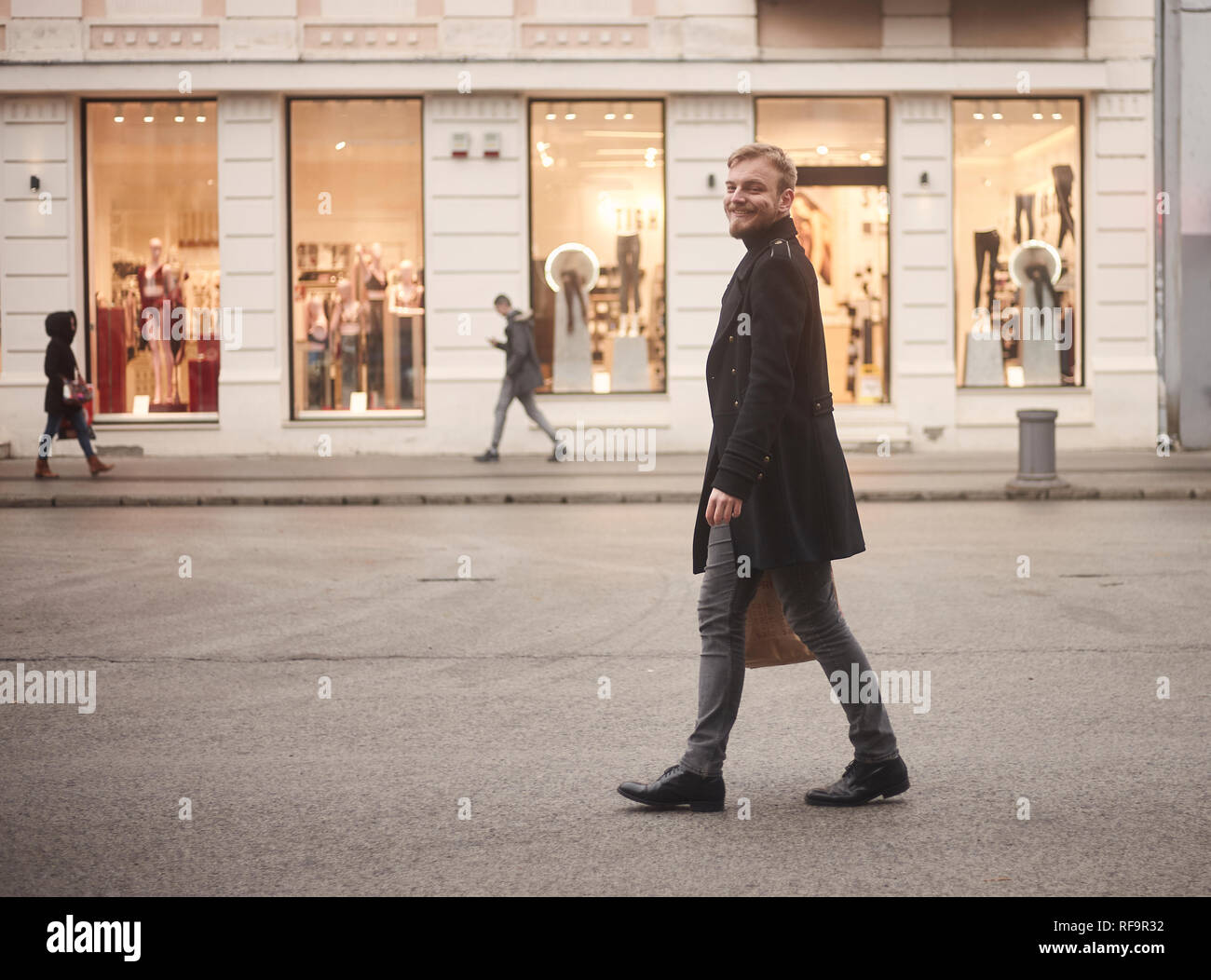Un joven sonriente y feliz el hombre, 20-29 años de edad, guapo y elegante, caminando por las calles de la ciudad sin coches, peatones caminando en segundo plano con st Foto de stock