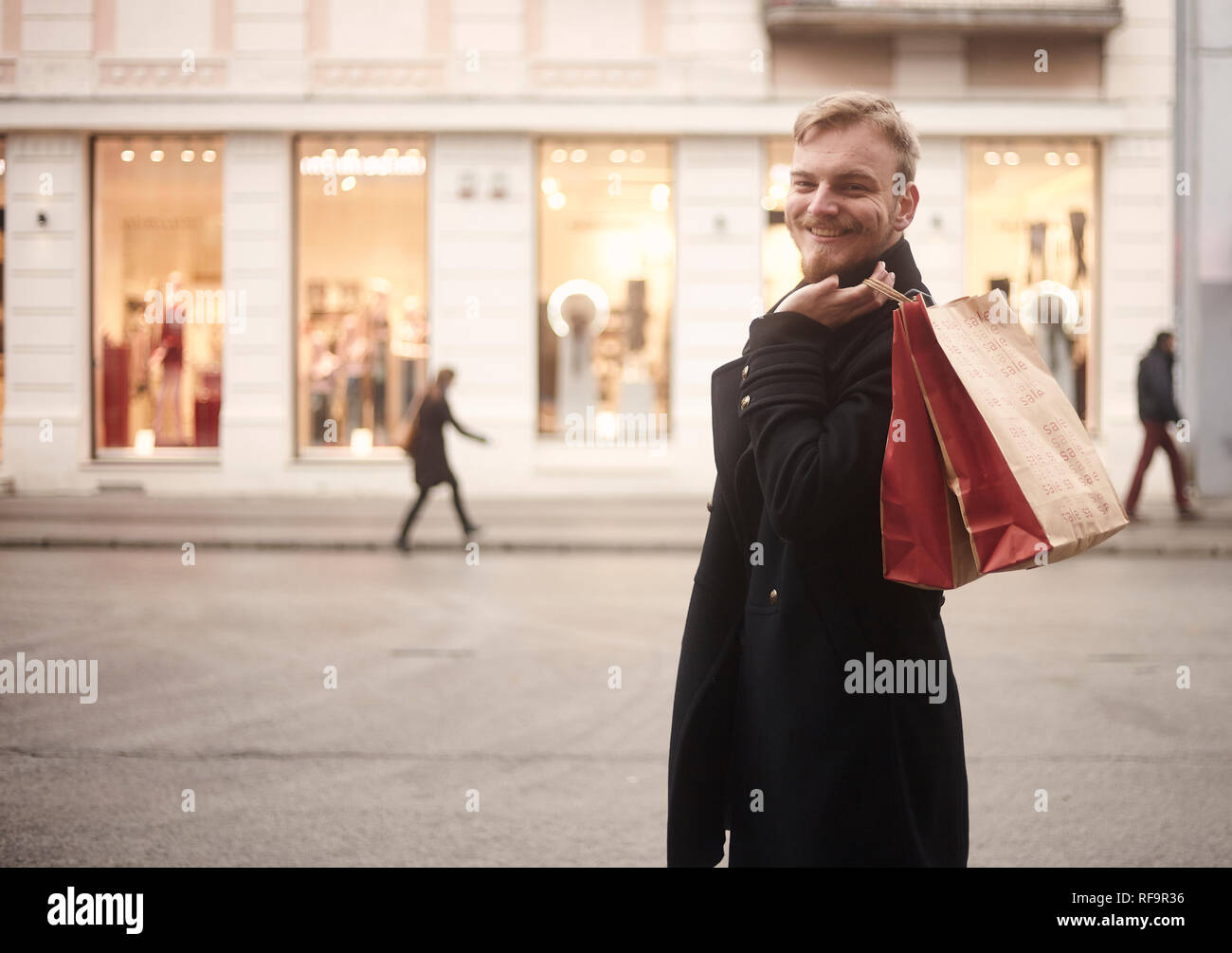 Un joven sonriente y feliz el hombre, 20-29 años de edad, guapo y elegante, mirando a la cámara mientras mantiene dos bolsas de compras sobre su espalda. Los peatones wal Foto de stock