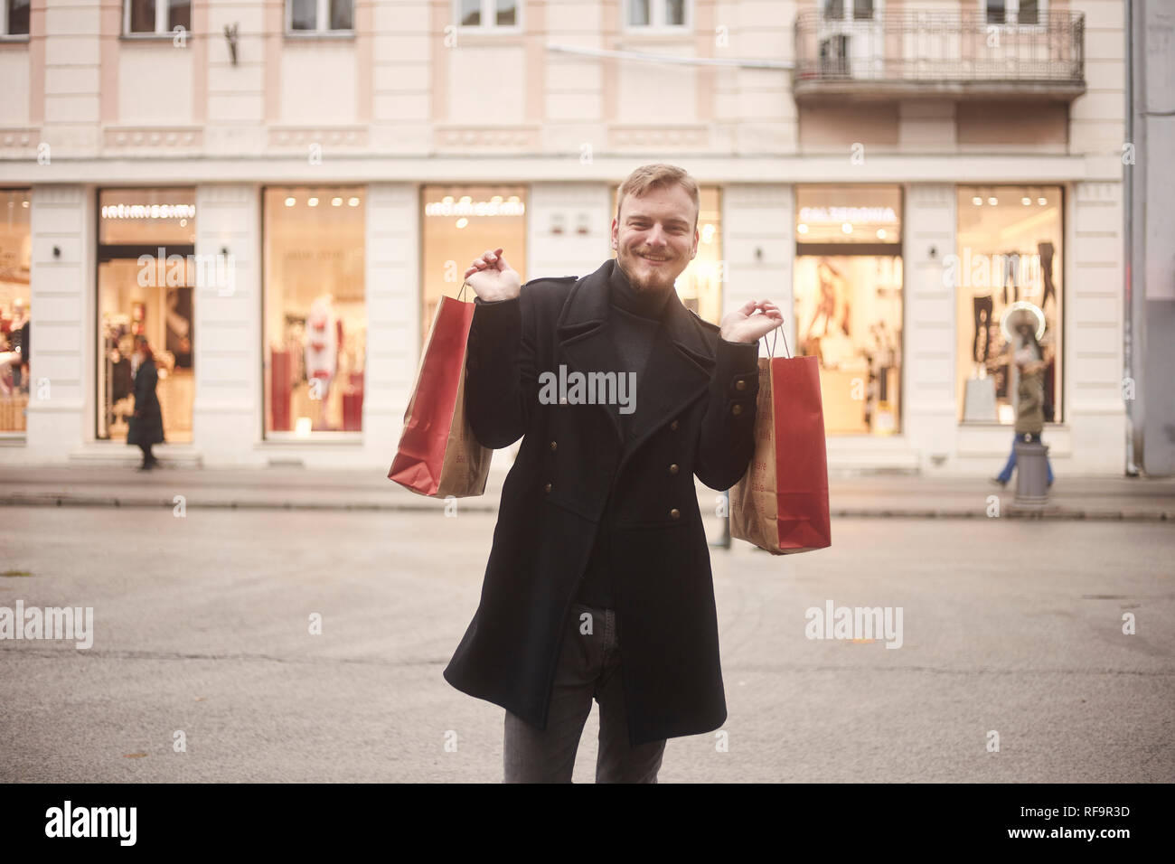 Un joven sonriente y feliz el hombre, 20-29 años de edad, en una calle peatonal, mirando a la cámara mientras mantiene dos bolsas de la compra con los brazos extendidos. S Foto de stock