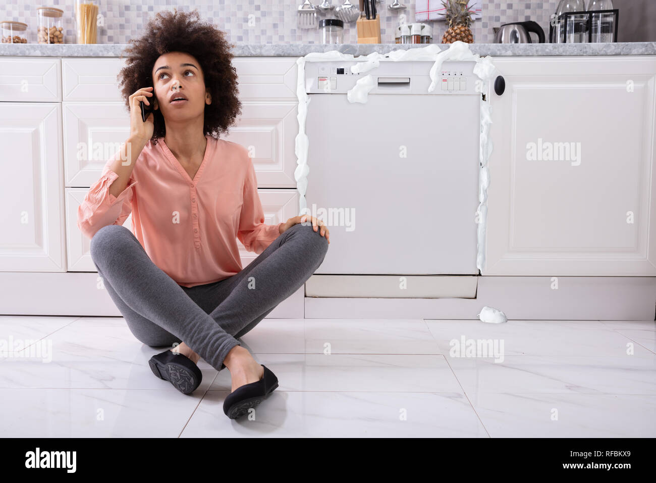 Triste Mujer llamar a soporte técnico por teléfono celular para arreglar  lavavajillas con espuma saliendo de ella Fotografía de stock - Alamy