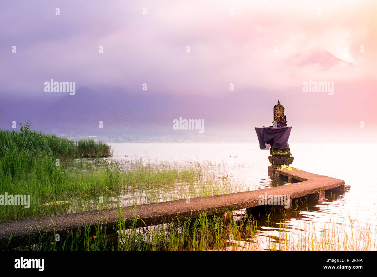 Pequeño templo hindú en el Lago con el puente de conexión durante el amanecer en Bali el día nublado y brumoso con la montaña como telón de fondo. Foto de stock
