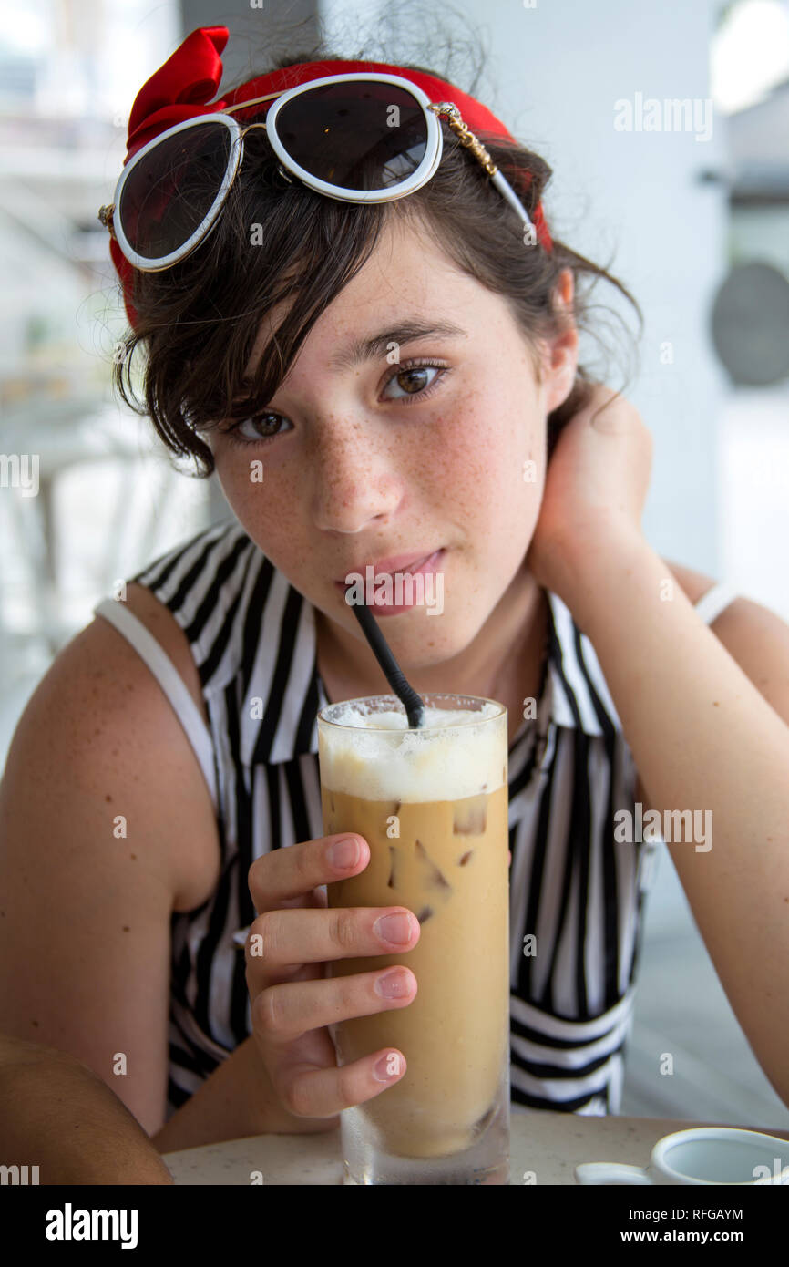 Una adolescente con bastante pecas en su rostro, vestida con una blusa de verano y con gafas de sol sobre su cabeza, bebidas café helado a través de una pajita en un c Foto de stock
