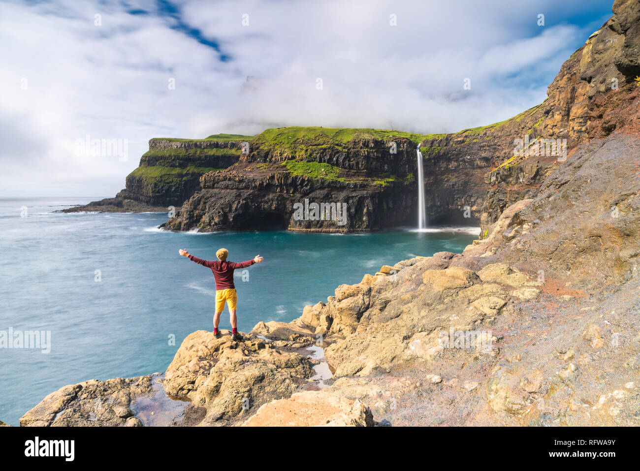 El hombre en los acantilados con los brazos abiertos, admirando Gasadalur cascada, isla de Vagar, Islas Feroe, Dinamarca, Europa Foto de stock