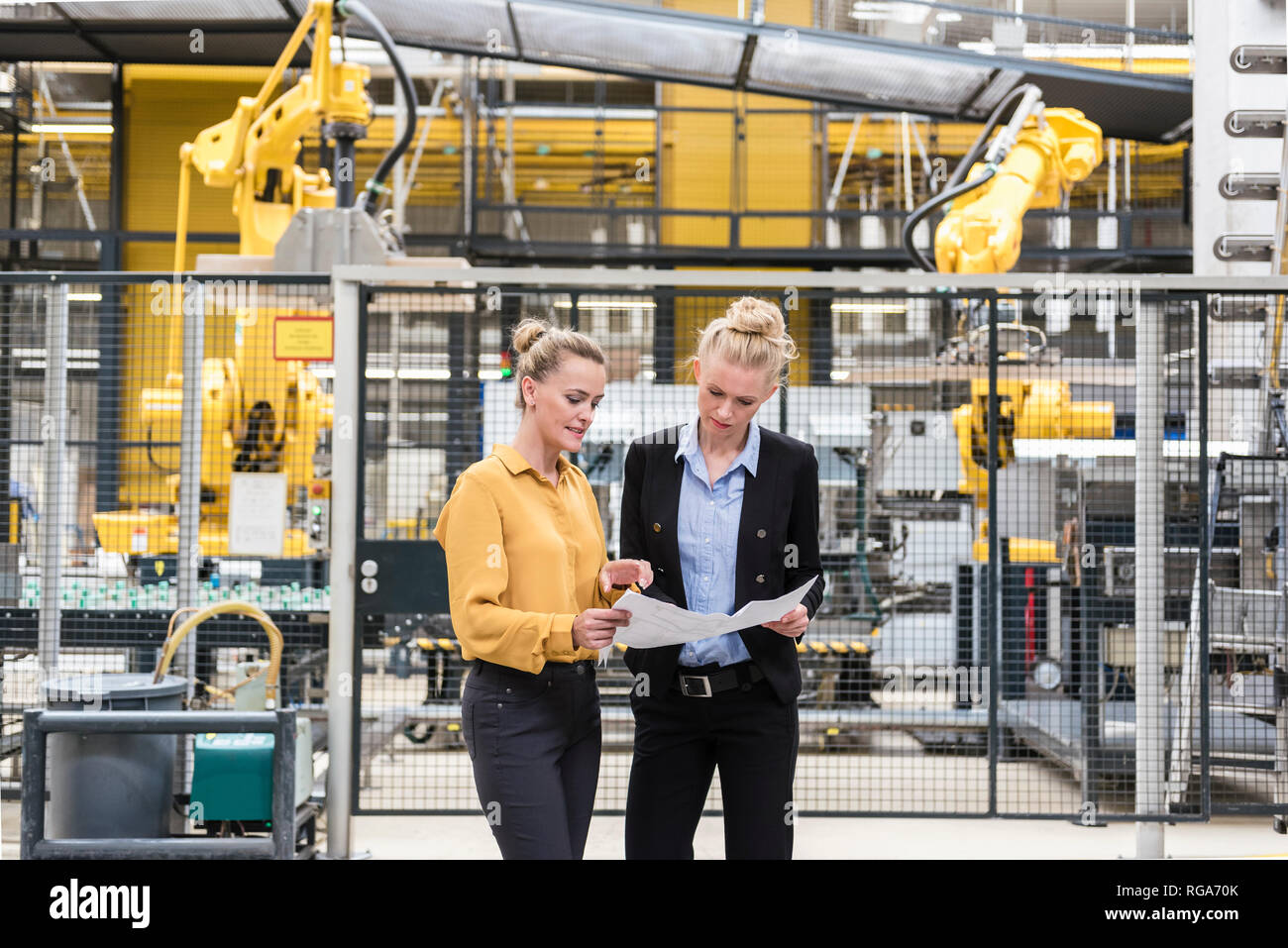 Dos mujeres hablando de plan de planta de fabricación en fábrica con robot industrial Foto de stock