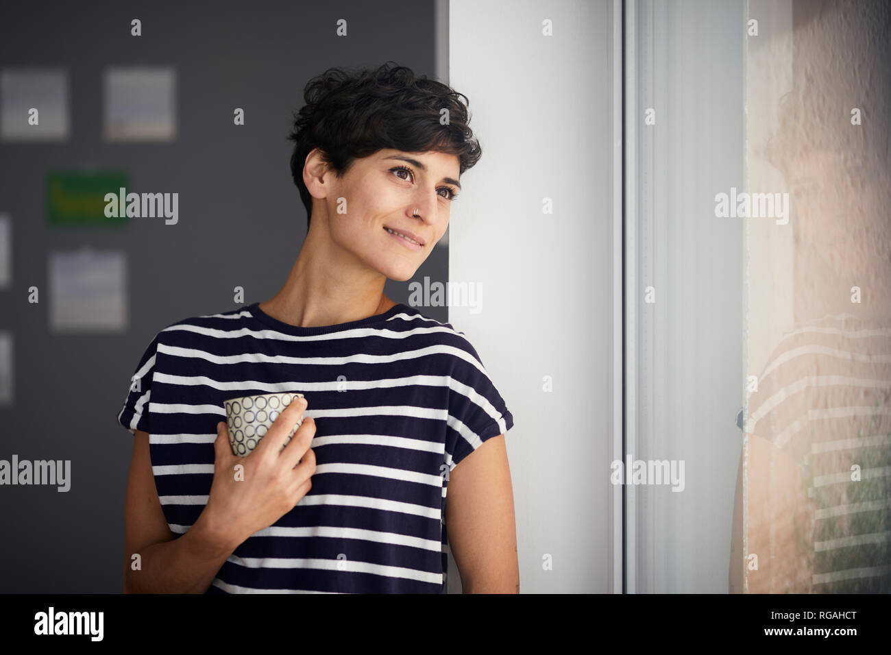 Mujer sonriente con una taza de café mirando hacia afuera de la ventana Foto de stock