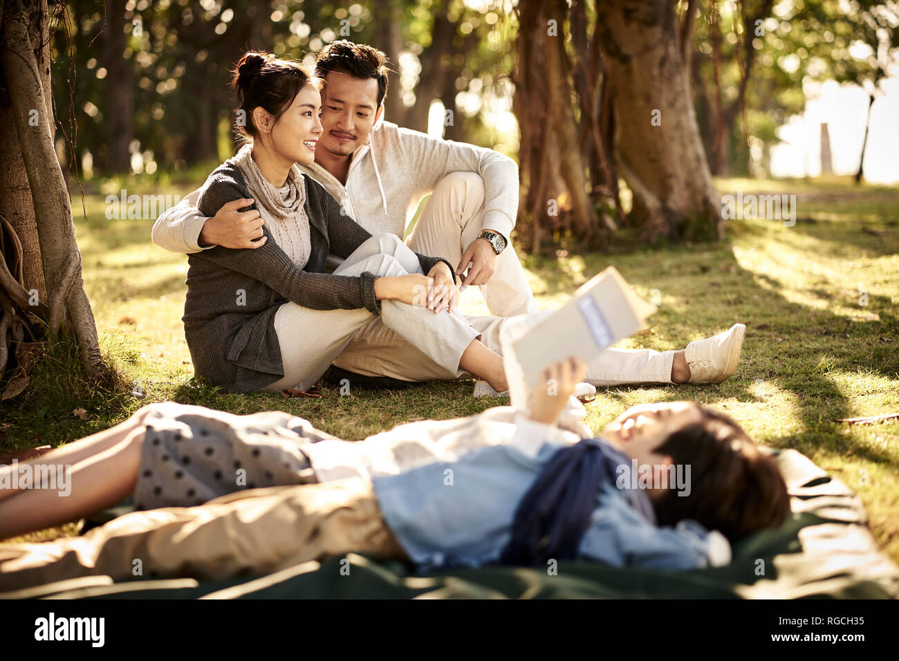 Pareja joven asiático sentados sobre el césped en el parque conversando con dos niños mentir libro de lectura en primer plano. Foto de stock