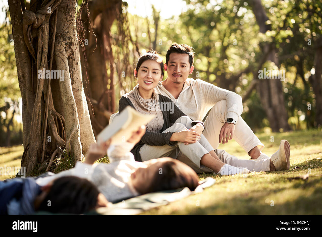 Dos niños asiáticos chico y chica divirtiéndose tumbado sobre el césped a leer un libro con sus padres sentados mirando en el fondo, se centran en los padres en b Foto de stock