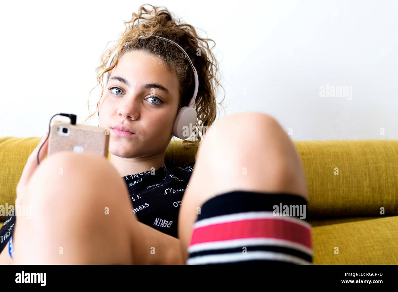 Retrato de muchacha sentada en el sofá escuchando música con auriculares y smartphone Foto de stock