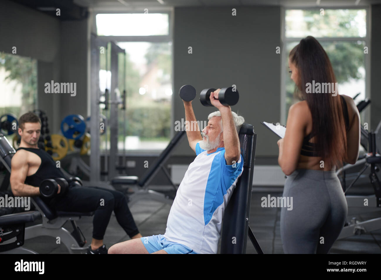 Closeup vista horizontal de 3 personas en el gimnasio. Atractiva joven morenita en ropa deportiva y la formación permanente elderler hombre. Contentrated fuertes graves altos hombre sentado y haciendo ejercicios weigt. Foto de stock