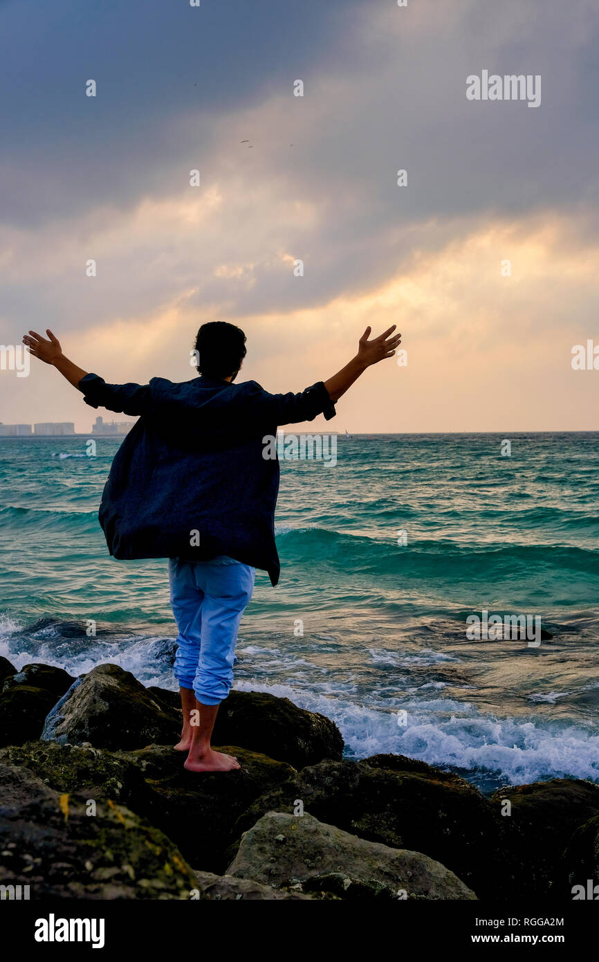 Un joven adulto de pie sobre las rocas en la playa con sus brazos abiertos amplia Foto de stock