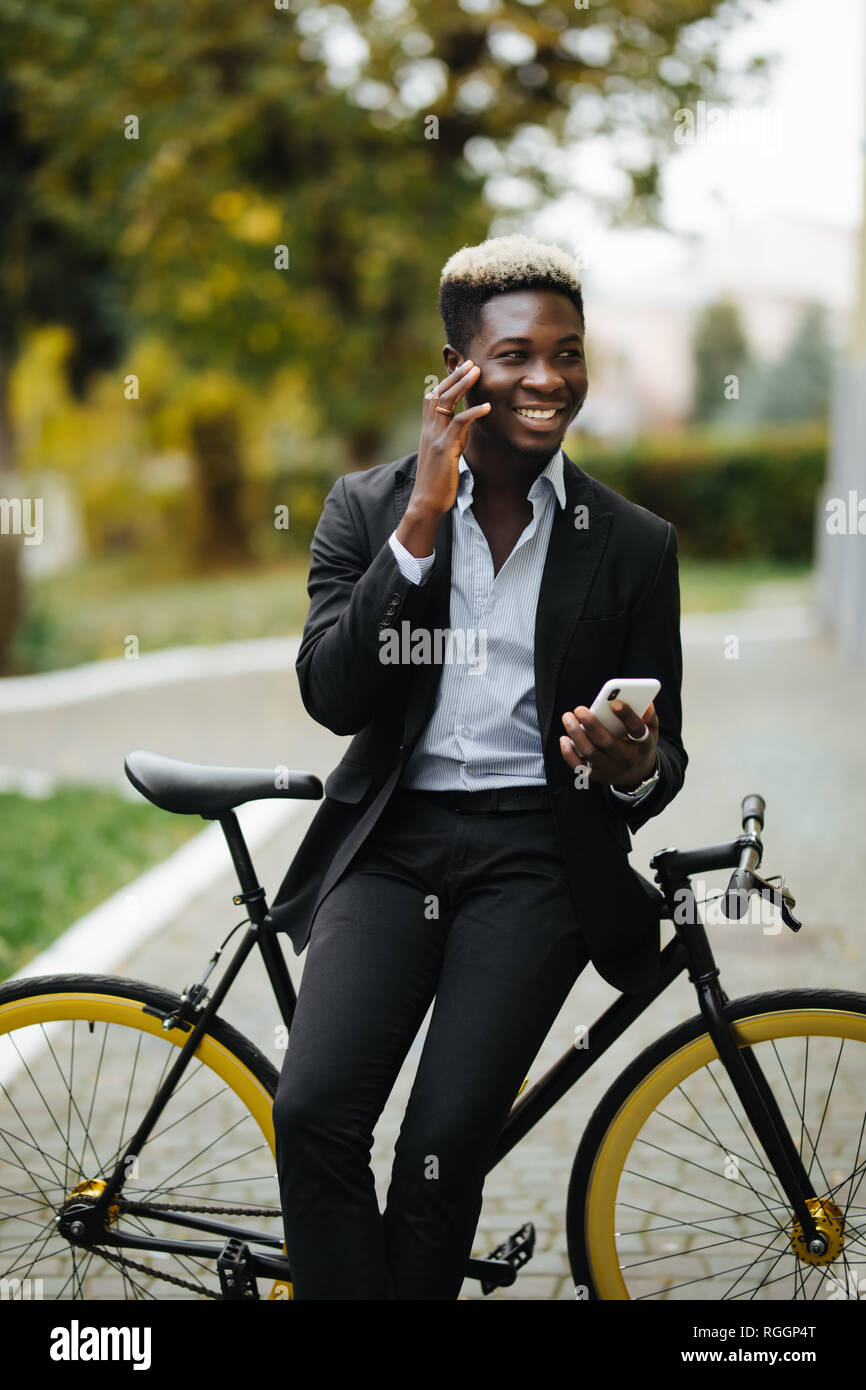 Apuesto joven hombre vestido con traje africano habla por teléfono mientras camina con bicicleta al trabajo por la mañana. El empresario va a trabajar con su bicicleta. Foto de stock