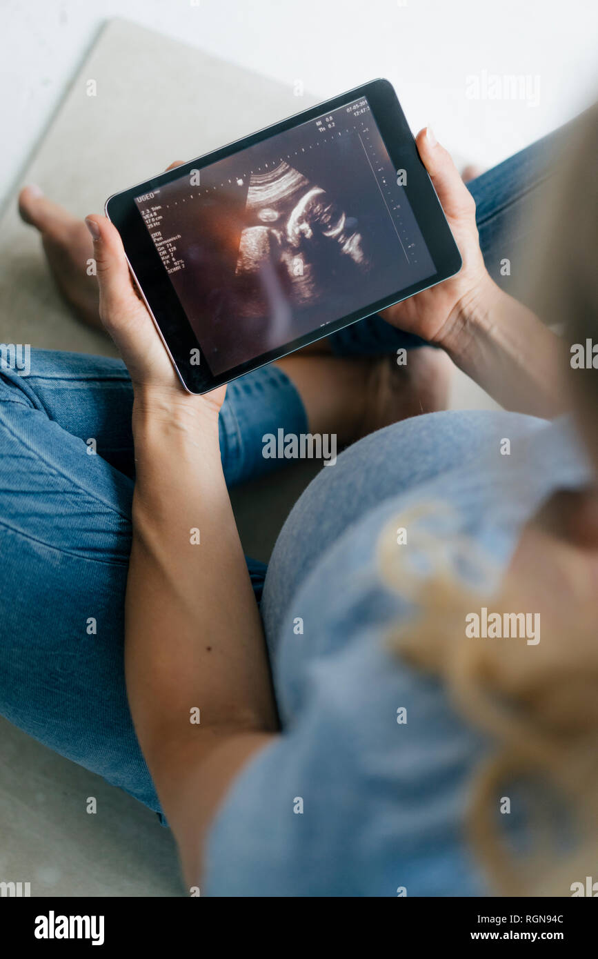 Mujer embarazada sentada en el suelo mirando la imagen de ultrasonido en tablet pc Foto de stock