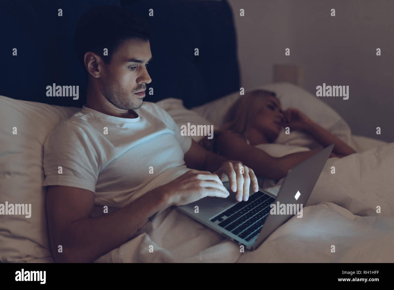 Joven sospechoso mediante ordenador portátil mientras novia dormir en la cama en la noche Foto de stock