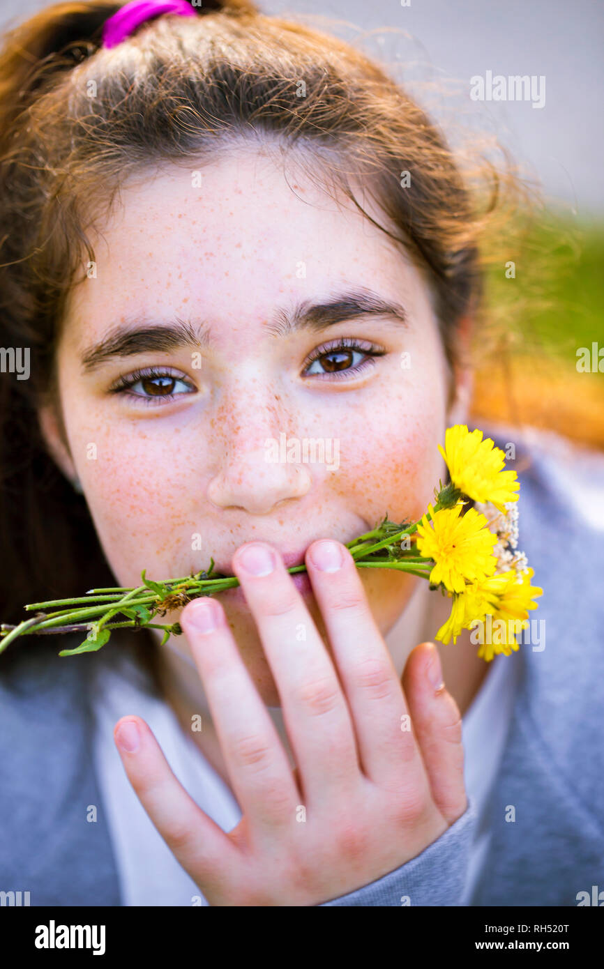 Close Up retrato de un alegre jovencita con largo pelo castaño travesuras y celebración amarillo el diente de león en su boca. Ella es lindo y divertido. Foto de stock