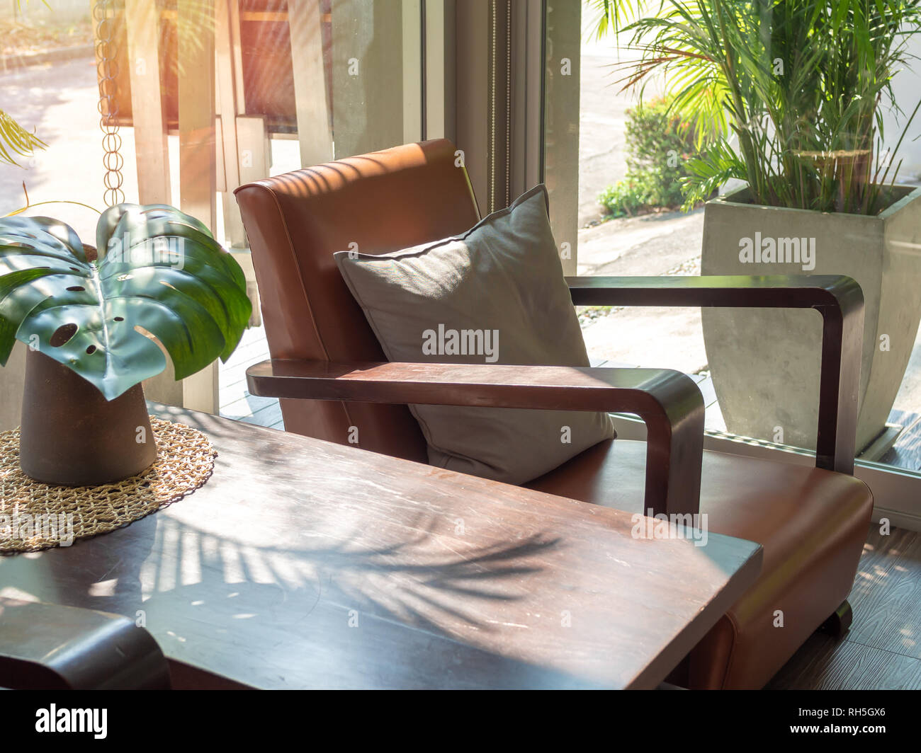 Sofá estilo retro de madera marrón con gris almohada y mesa de madera con  hojas de palmeras verdes en un jarrón cerca de la ventana de vidrio.  Sillones de cuero en la