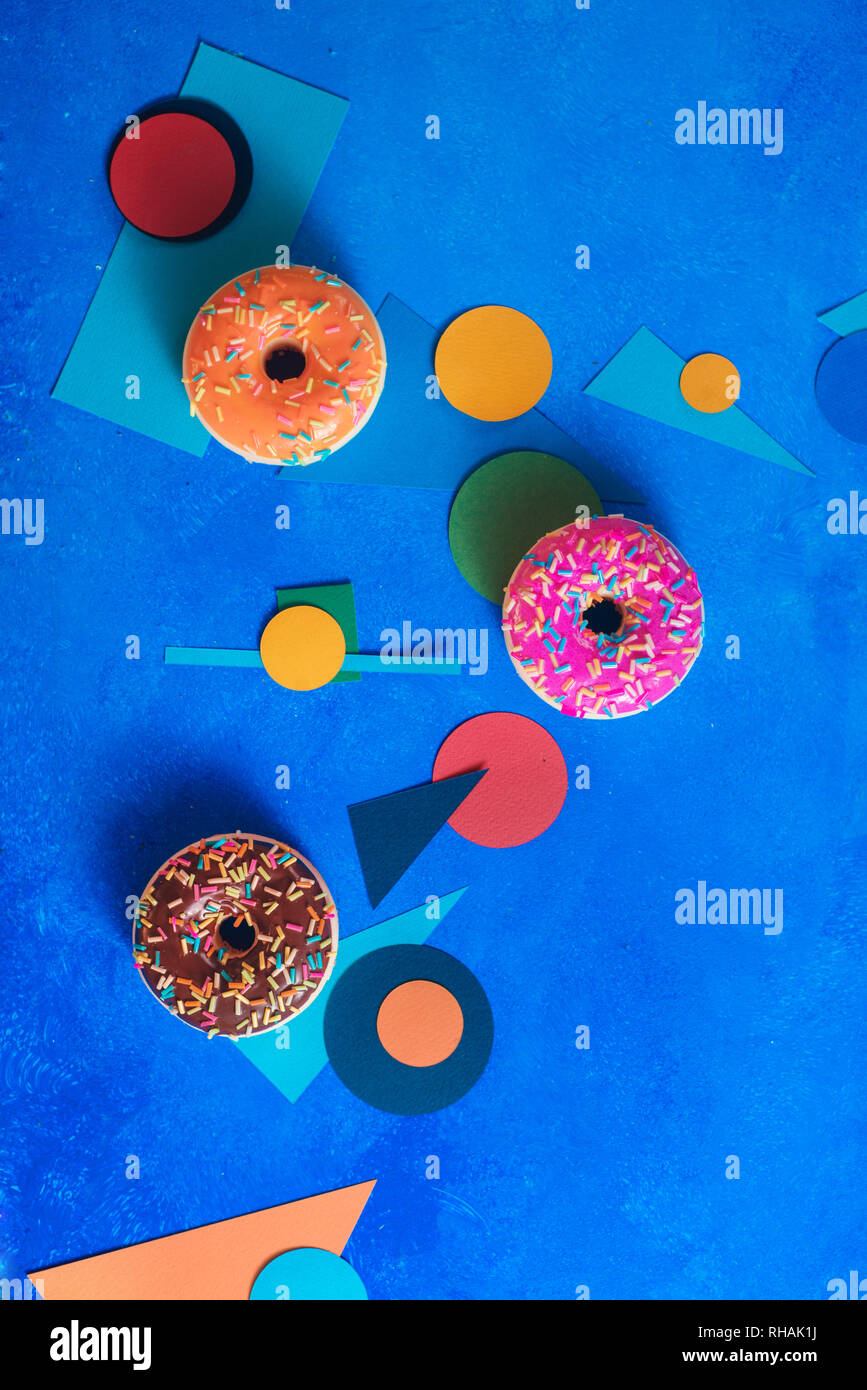 Buñuelos de diferentes colores sobre un fondo azul con formas geométricas. El bloque de color plano de alimentos se acostó con espacio de copia. Foto de stock