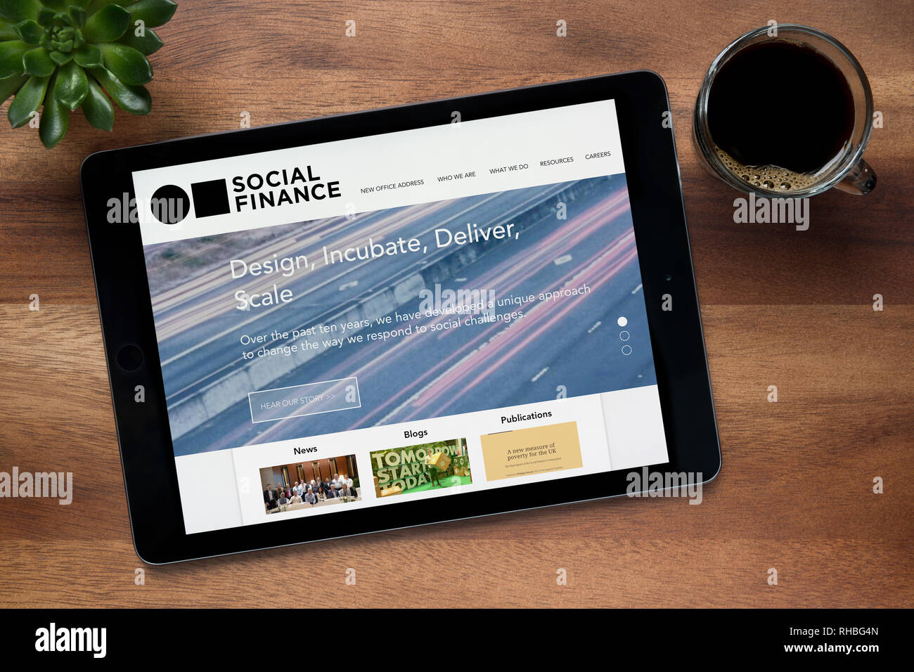 El sitio web de la financiación social es visto en un iPad, en una mesa de madera junto con un café espresso y una planta de casa (Editorial). Foto de stock
