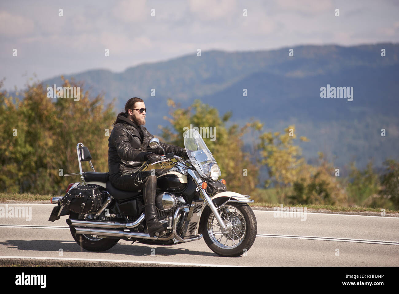 Vista lateral del barbudo motociclista de pelo largo en gafas de sol y ropa de cuero negro caballo cruiser moto a lo largo de angostas camino de asfalto en día soleado Foto de stock