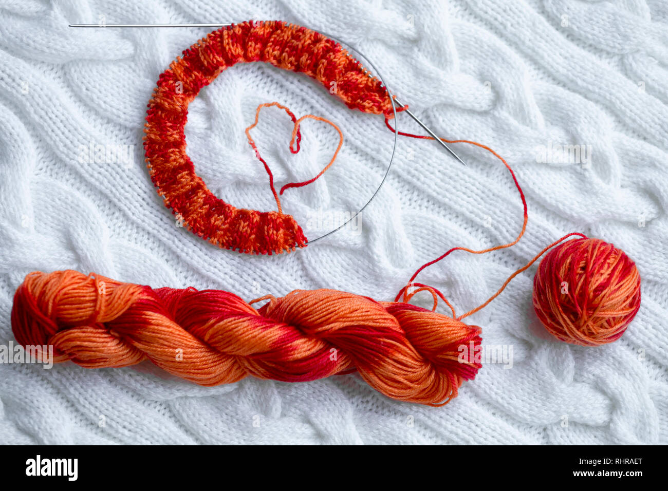 Proyecto de tejer en progreso. Un trozo de tejido con una bola y una madeja de color rojo-naranja y agujas de tejer contra un blanco tejida de cuadros escoceses. Foto de stock