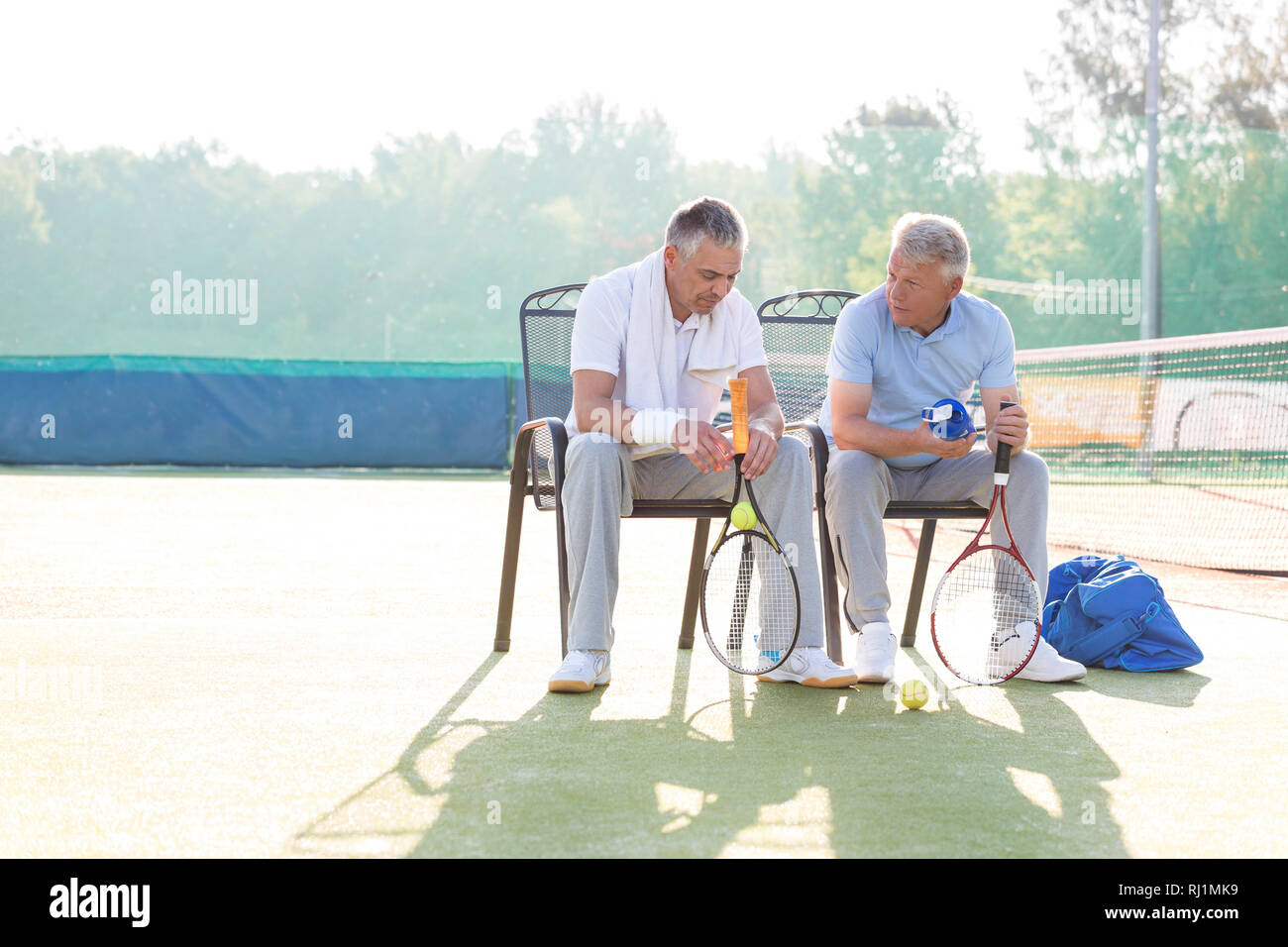 La longitud total del macho maduro amigos hablando mientras sentado con raquetas y pelotas de tenis en silla en la corte Foto de stock