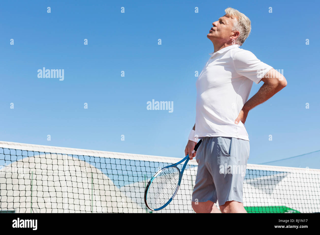 Hombre maduro gesticulaciones con dolor de espalda sujetando raqueta de tenis contra el azul claro del cielo en día soleado Foto de stock