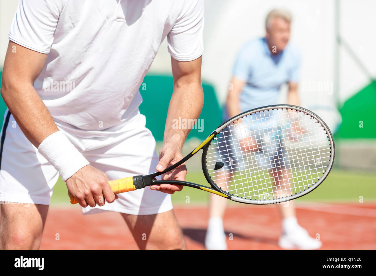 La mitad del torso de hombre de pie con raqueta de tenis contra un amigo jugando partido de dobles en la cancha Foto de stock