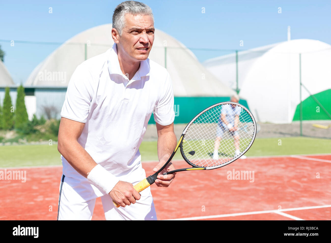 Seguros de hombre maduro de pie con raqueta de tenis contra un amigo jugando partido de dobles en la cancha Foto de stock
