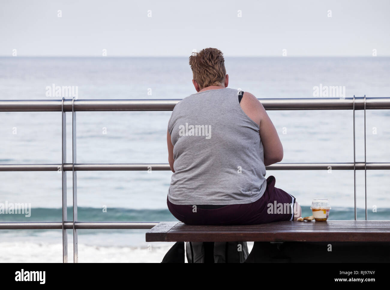 Vista trasera de la obesa mujer con bocadillos azucarados sentado en un banco con vistas al mar. Foto de stock