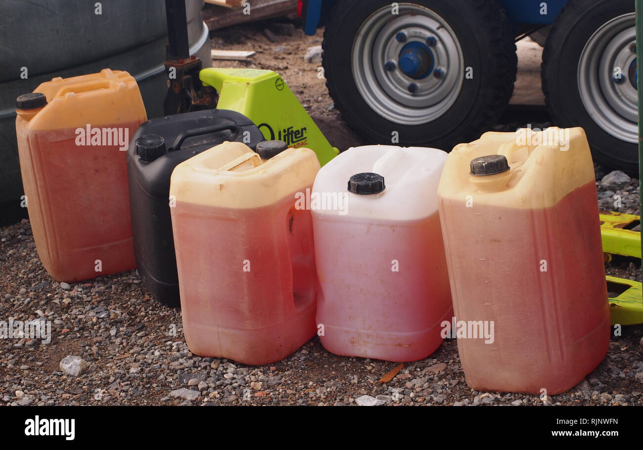Una línea de galón plástico recipientes que contengan líquidos industriales frente a los vehículos de ruedas y brazos de levantamiento en una pista de grava. Foto de stock