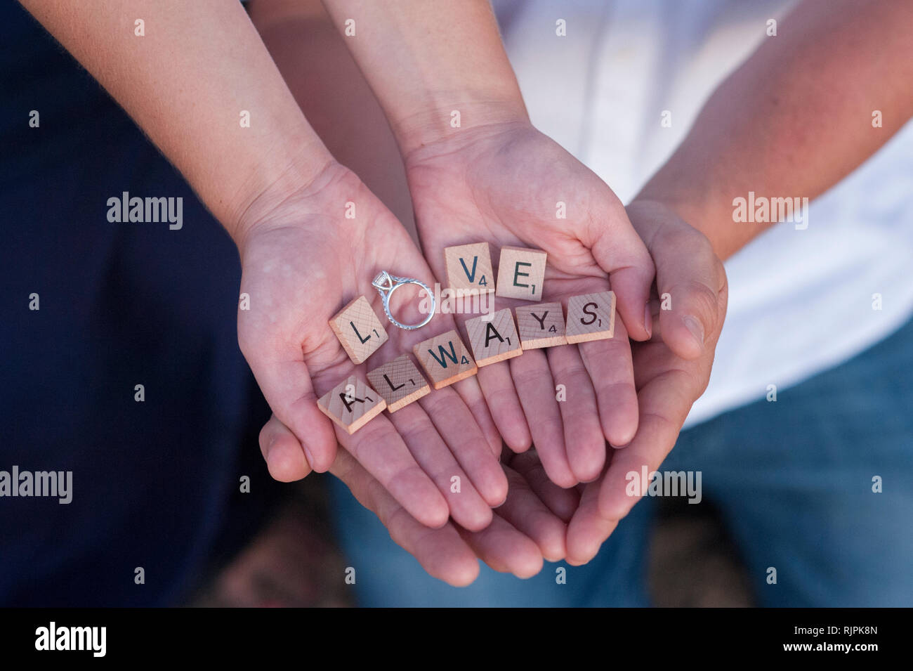 Cierre horizontal de las manos de la pareja ahuecada alrededor de cada una de las demás piezas de Scrabble, sujetando la ortografía de las palabras "el amor siempre' con el anillo. Foto de stock