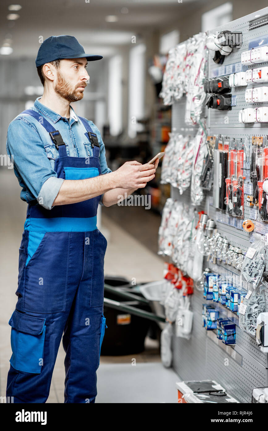 Guapo electricista en ropa de trabajo eligiendo artículos pie con teléfono inteligente en el edificio shop Fotografía stock - Alamy