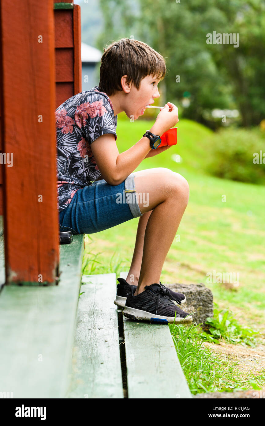 Retrato de muchacho sentado fuera de cabaña de madera, comer el desayuno. Flam, Noruega, Europa Foto de stock
