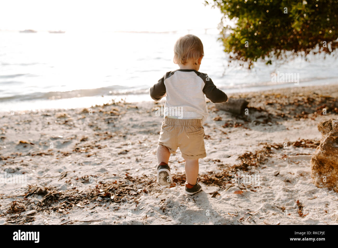 Precioso lindo Adorably Happy Baby Boy niño sonriente y jugando en la arena de la playa al lado del agua del océano fuera durante la puesta de sol en el verano Foto de stock