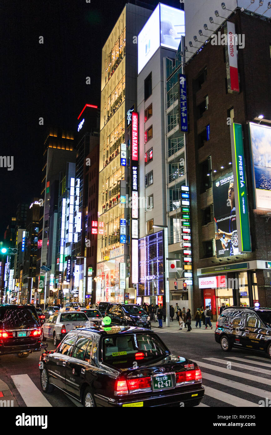 Ginza de Tokio, la noche. Insignia de la fachada de vidrio de 12 pisos Uniqlo almacenar entre otros almacenes de gran altura a lo largo de la calle. Taxi en primer plano. Foto de stock