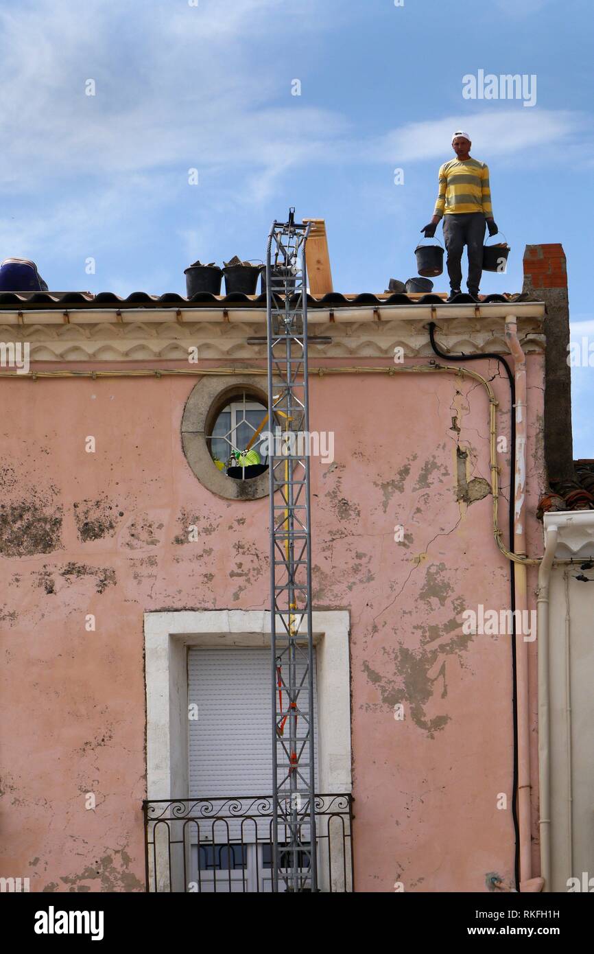 Un techador trabajando sobre un techo en el meze, Francce usa una cuchara escalera para tomar el material fuera de la azotea (cara Photoshopped). Foto de stock