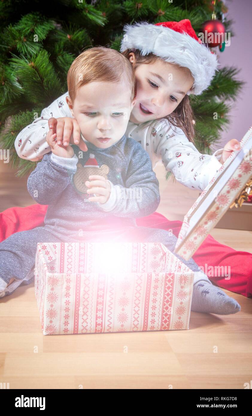 Hermanitos abriendo sus regalos cerca de árbol de Navidad. Son iluminados por la mágica luz de caja de regalo. Foto de stock