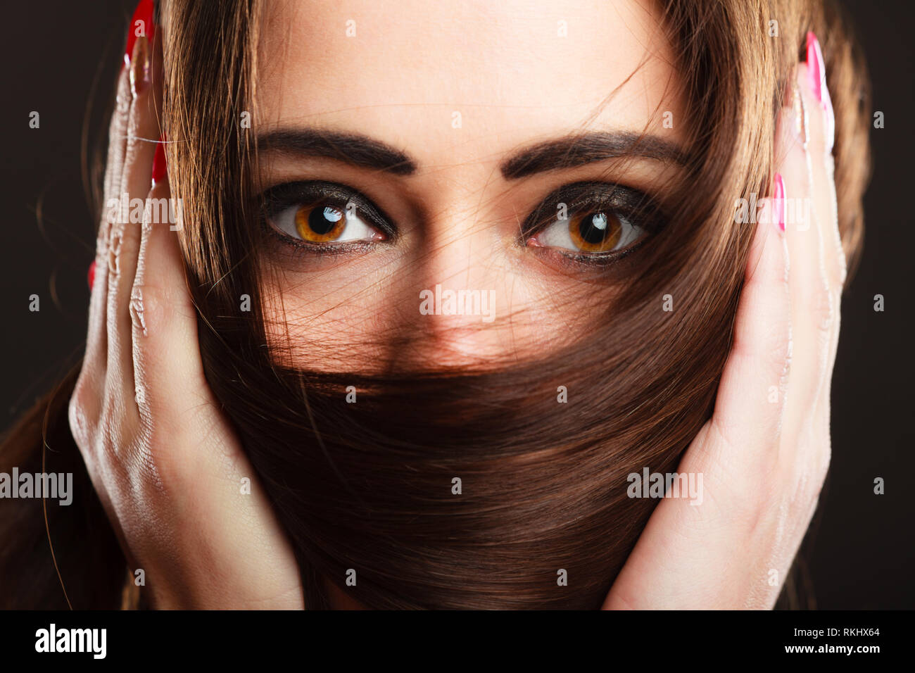 Cerrar mujer cubre el rostro por largos pelos marrones ojos marrones con maquillaje oscuro Foto de stock