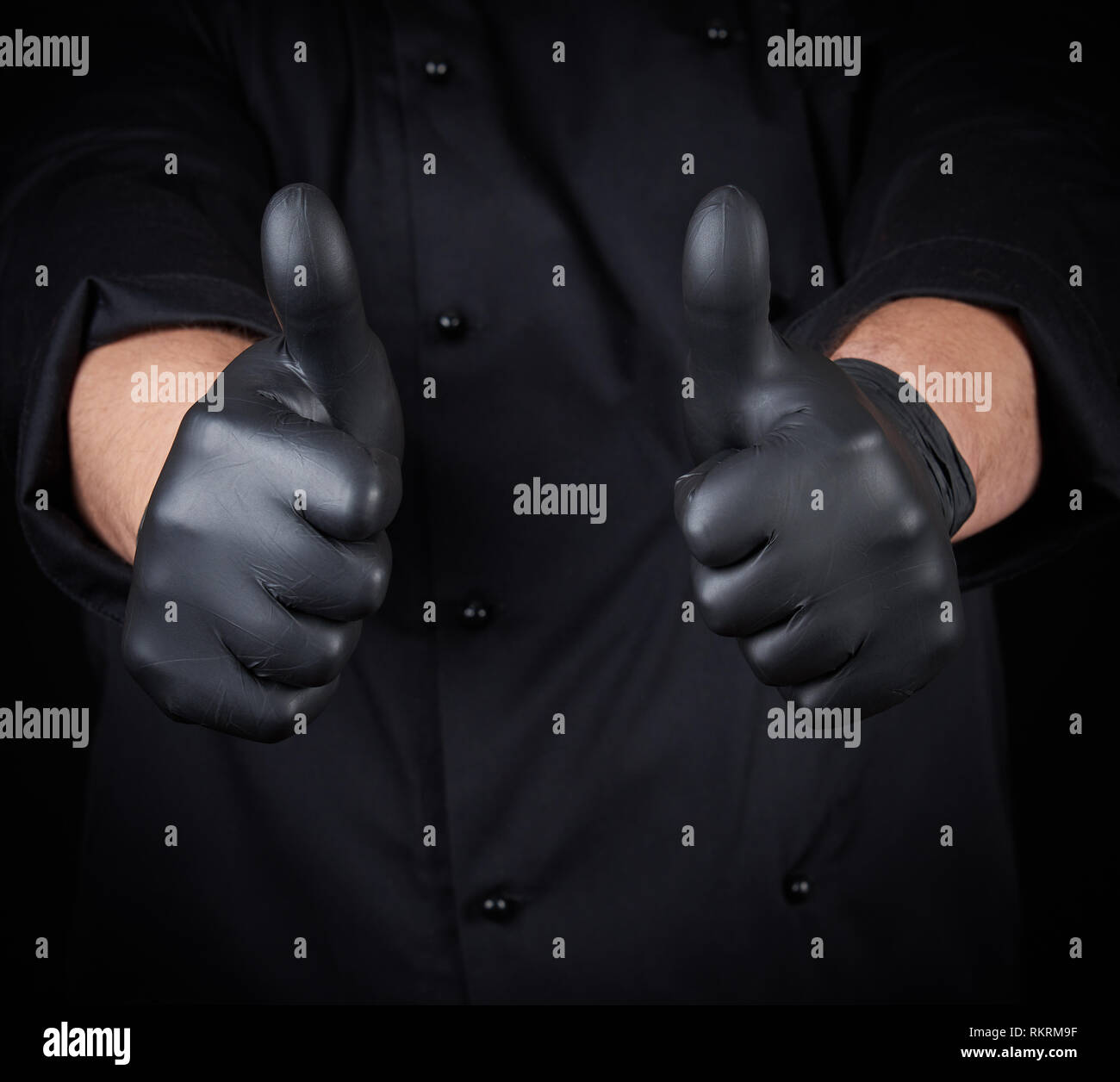 Macho de la mano del chef en negro guantes de látex y uniforme negro muestra un gesto de aprobación, como Foto de stock