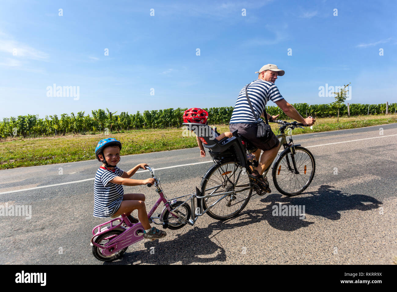 Paseo en bicicleta de la familia en el campo, padre con dos niños en casco en la carretera, asiento portabicicletas Foto de stock