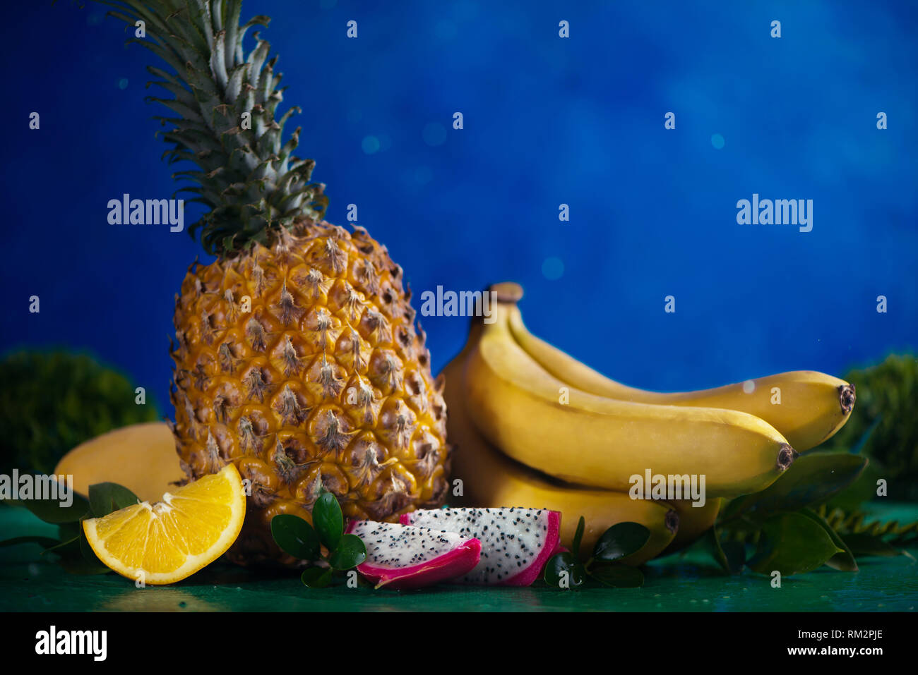 La piña, plátano, mango, dragon fruit con hojas tropicales sobre un fondo azul con gotas de agua y espacio de copia Foto de stock