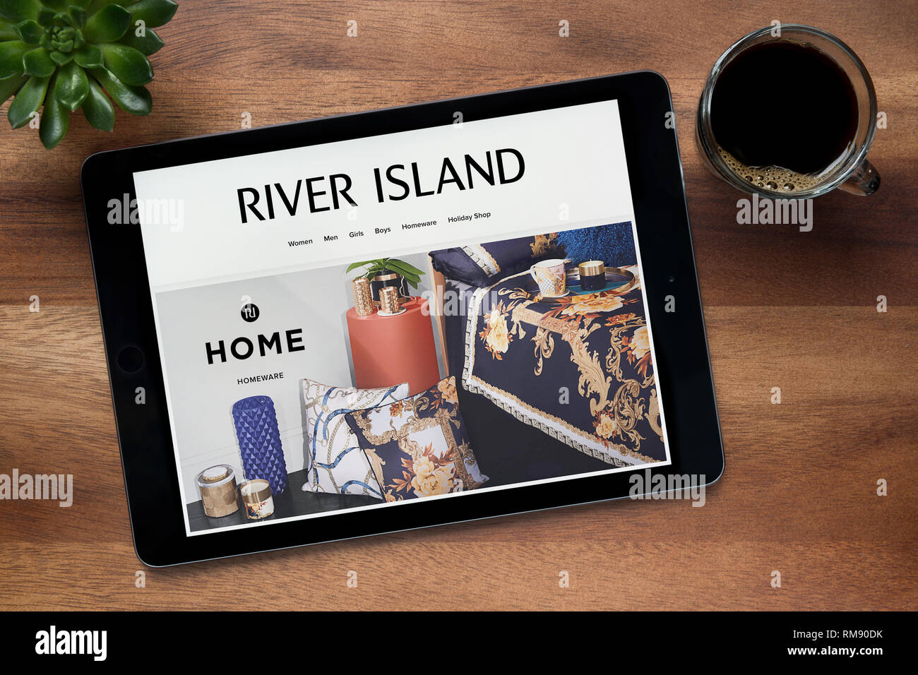 El sitio web de River Island es visto en un iPad, en una mesa de madera junto con un café espresso y una planta de casa (Editorial). Foto de stock