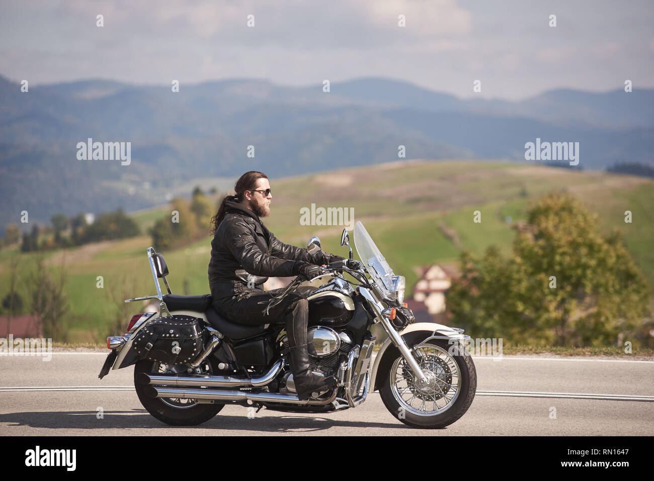 Vista lateral del barbudo motociclista de pelo largo en gafas de sol y ropa de cuero negro caballo cruiser moto a lo largo de angostas camino de asfalto en el soleado día de otoño. Foto de stock