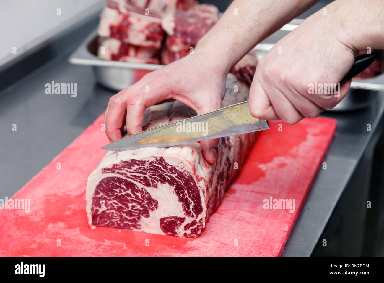 Primer plano de la mano de cocinar los cortes de carne de mármol chuletón  filete con un cuchillo afilado en la tabla de cortar de plástico rojo sobre  la mesa metálica en