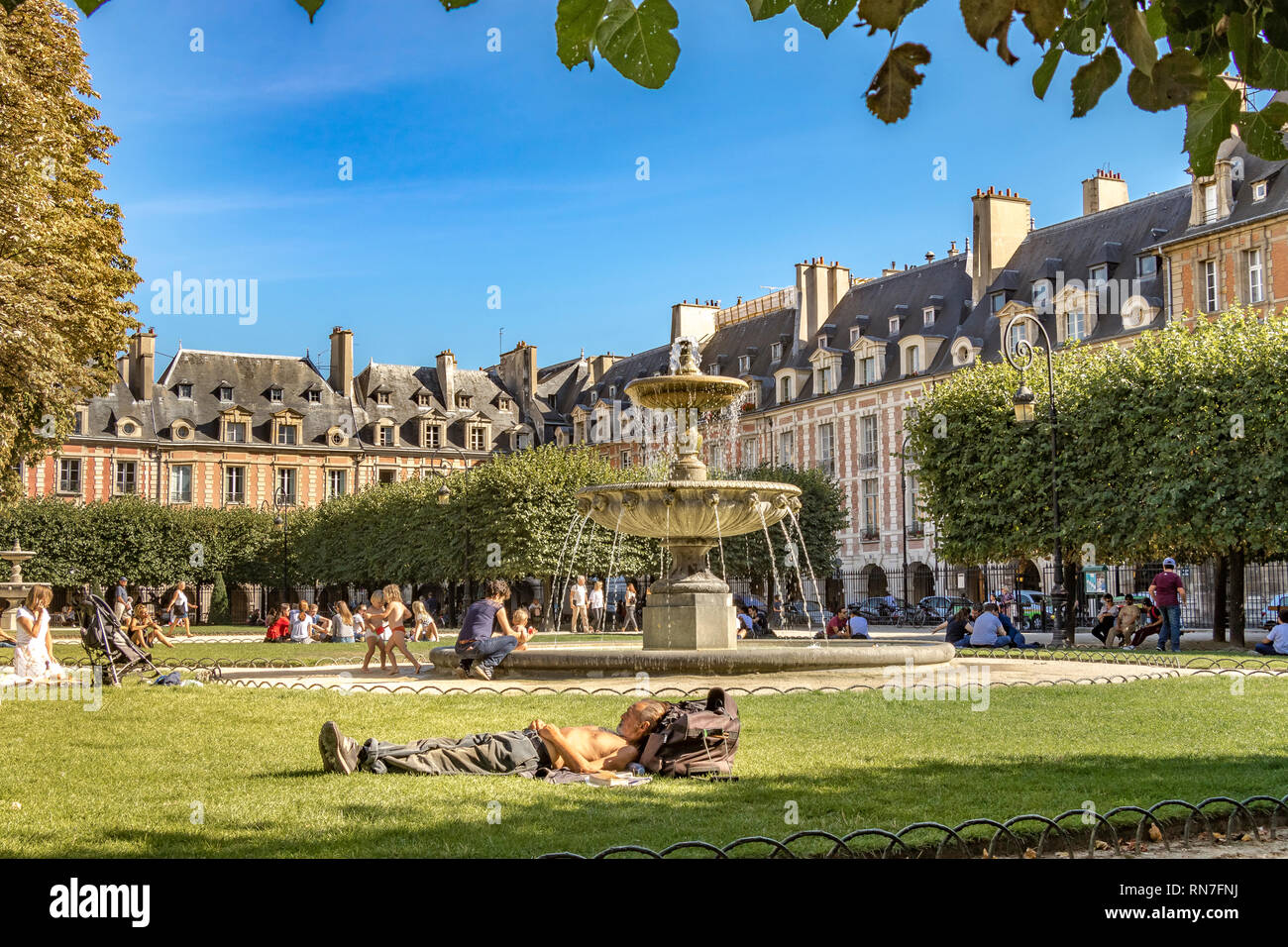 Gente sentada y tomando el sol en el césped cerca de la fuente en la Place des Vosges , en el distrito de moda Le Marais de París, Francia Foto de stock